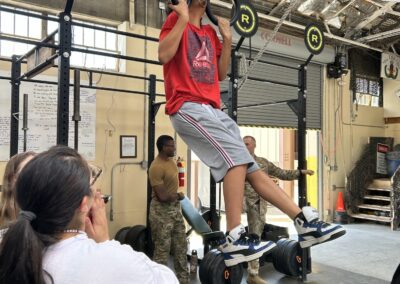 Youth performs a pull-up on gymnastics rings inside a military fitness facility while others watch